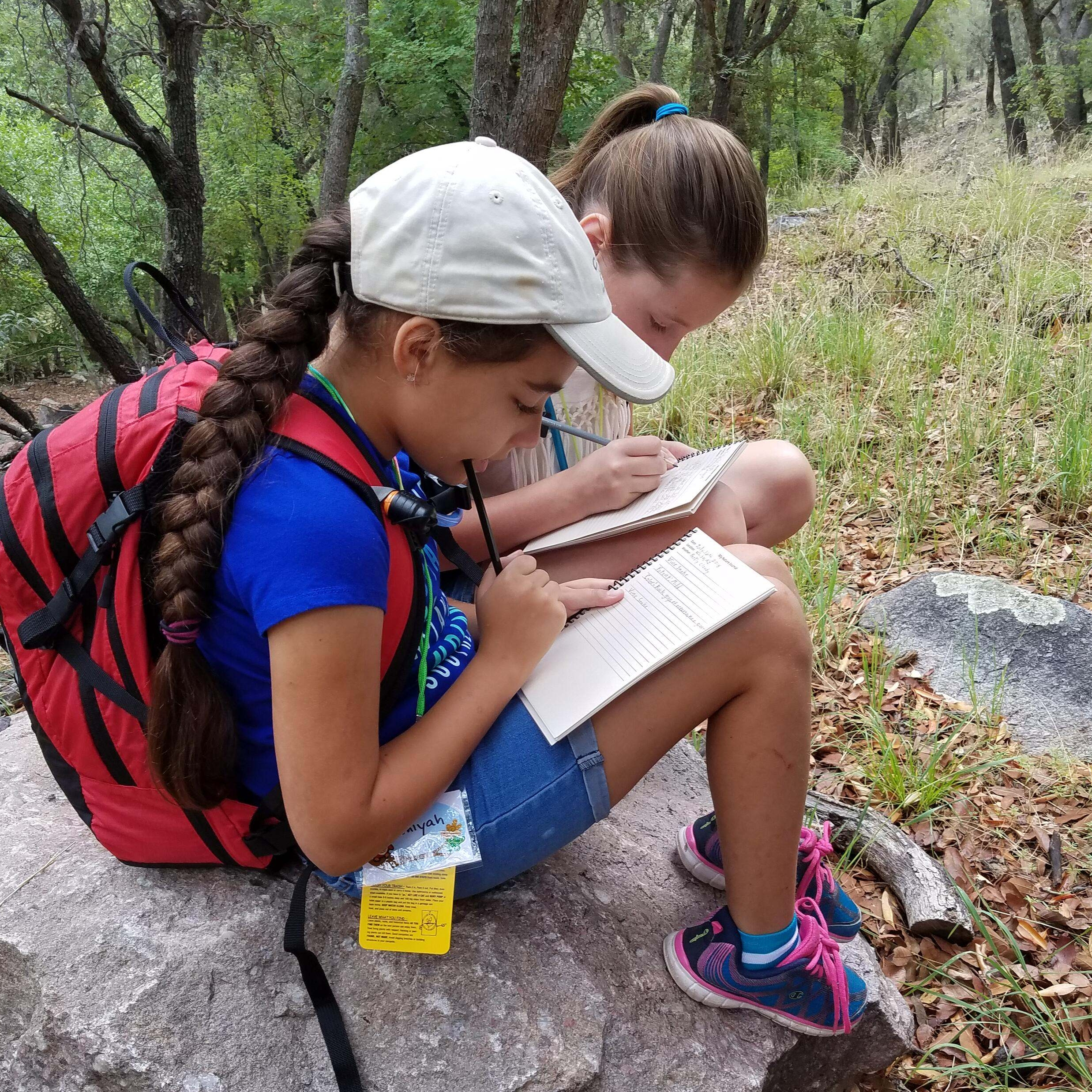 Two children, one wearing a backpack, sit on a rock and write in notebooks.