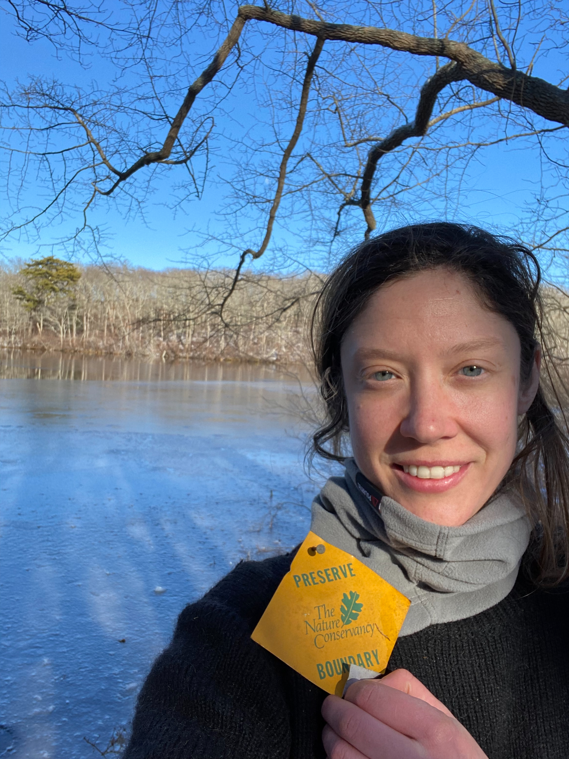 A selfie of Bekah Myers holding up a small TNC preserve boundary sign in front of a pond.