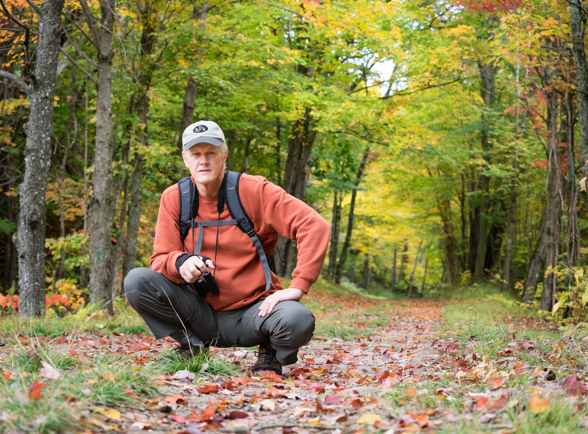 A person in outdoor gear crouches down and looks into the camera. 