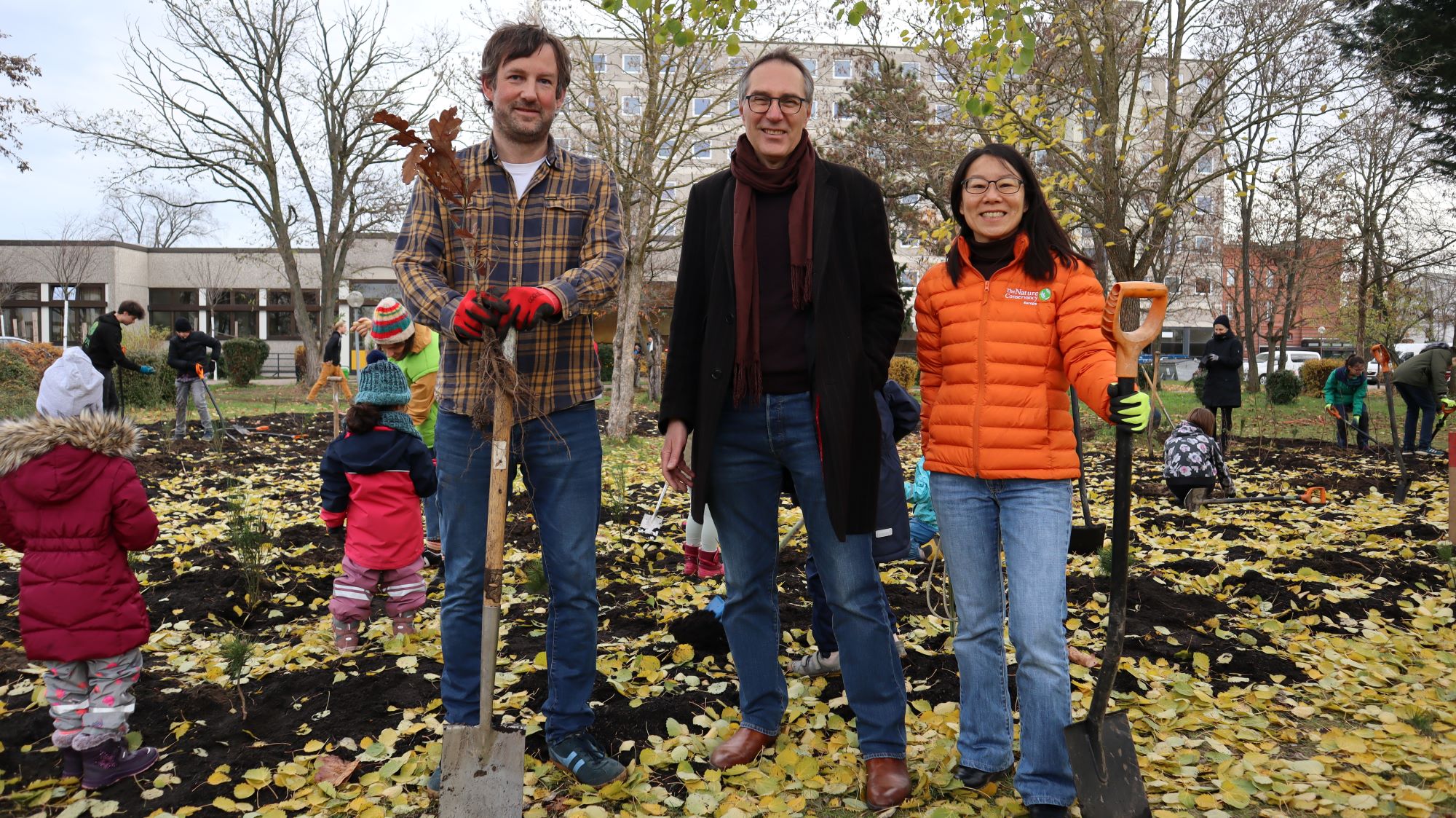 Three people standing in front of a tree planting even with little children running around.