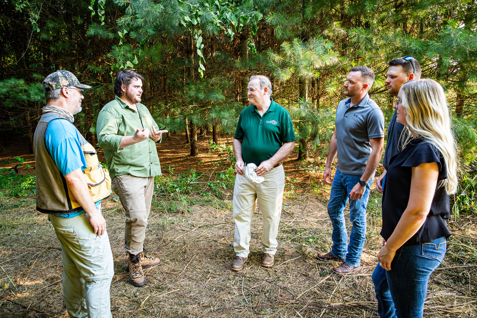 Six people stand in semi-circle facing one person who is talking in forested landscape.