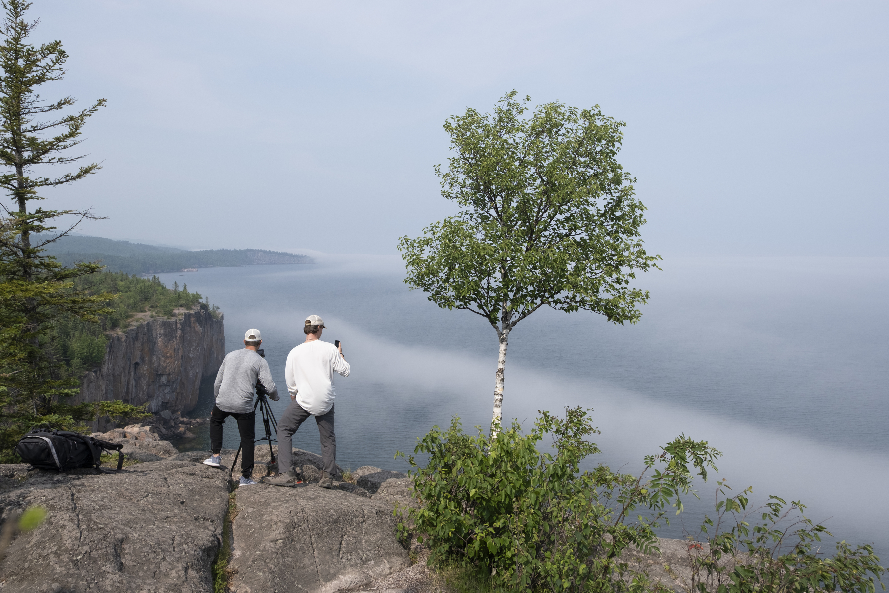 Two people stand next to a lone tree on a mountain overlook.