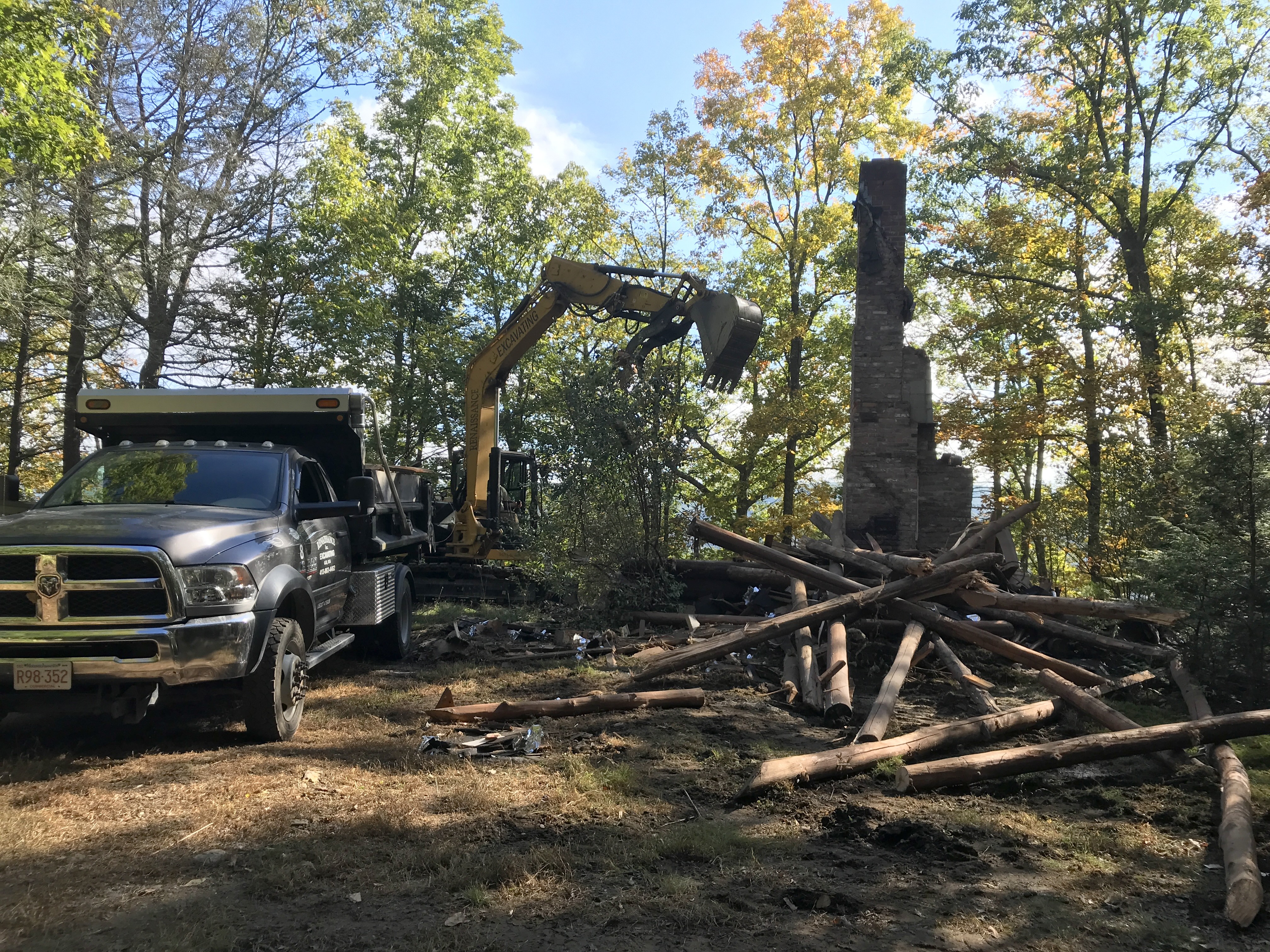 A bulldozer arm hovers over a pile of wood and a still-standing brick chimney, the remains of a wooden cabin. Autumnal trees are in the background.