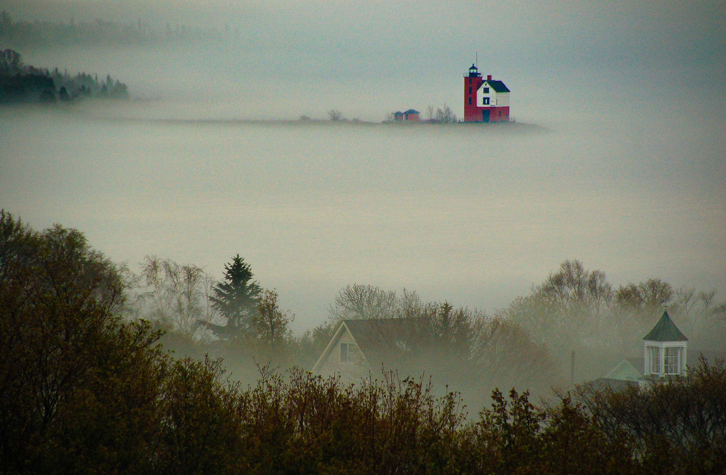 Mist covers a forest and homes. A red lighthouse is in the distance, partially covered in mist. 
