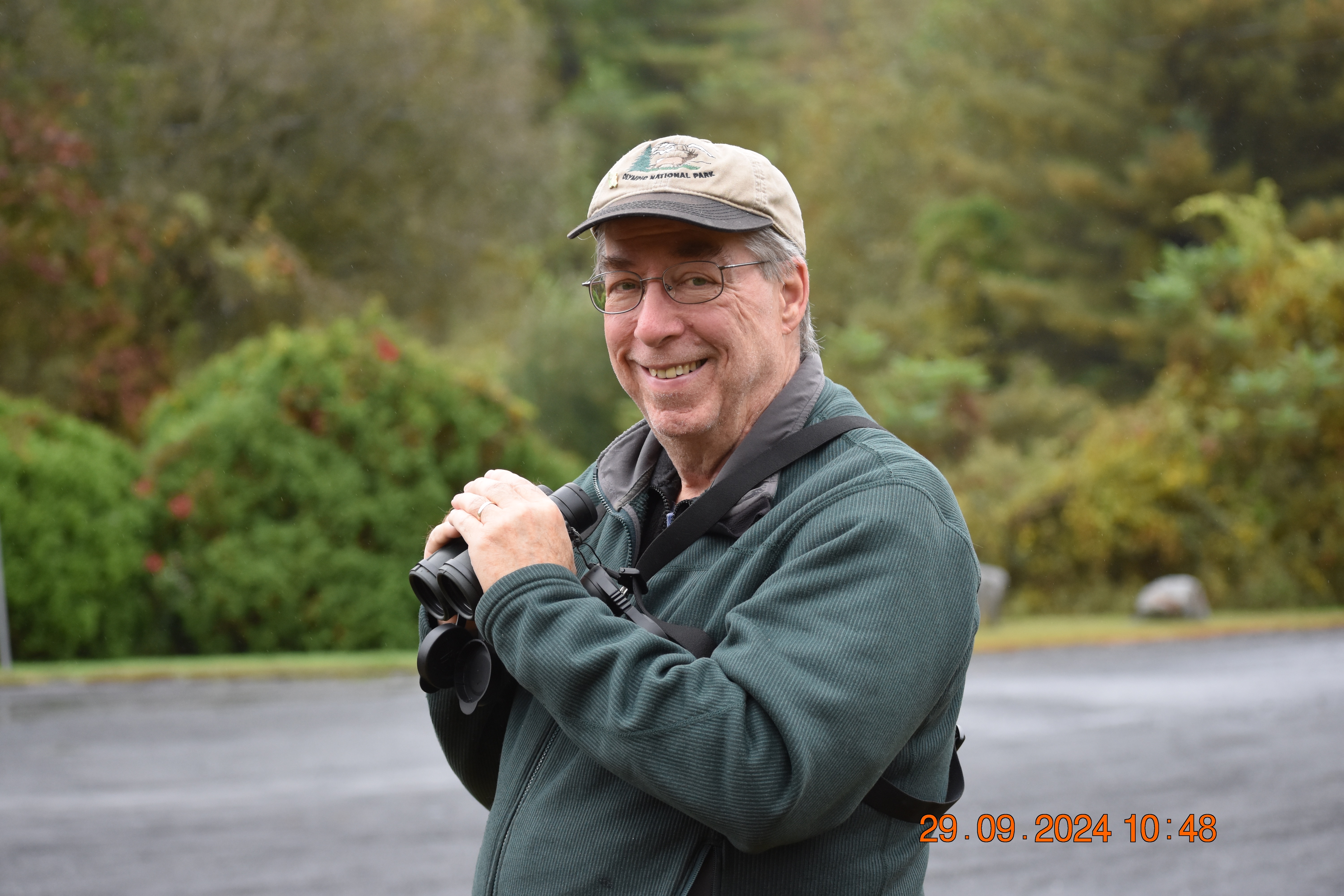 A person holding binoculars stands in the forest and looks at the camera.