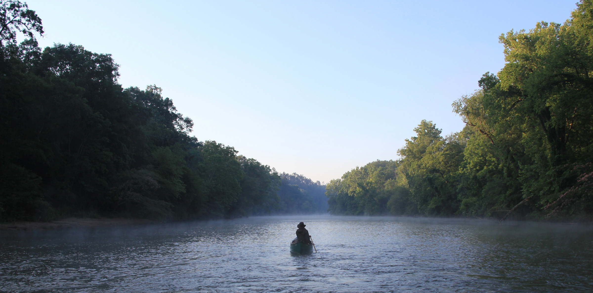 A kayaker floats on a calm river surrounded by forest.