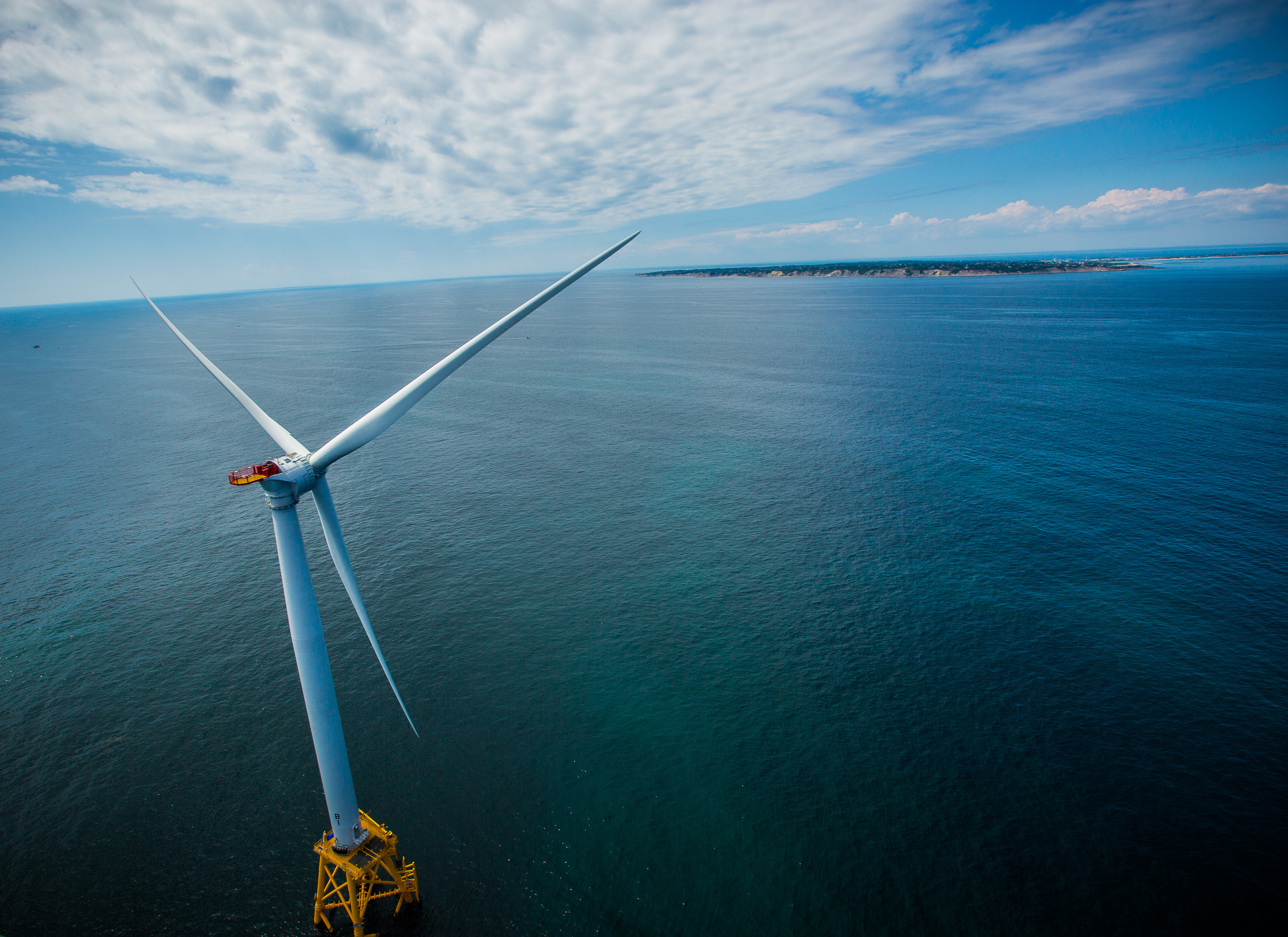 A wind turbine is pictured at left and sits on top of a wide open ocean, with a small patch of land with green grass in view. Clouds are seen in a blue sky.