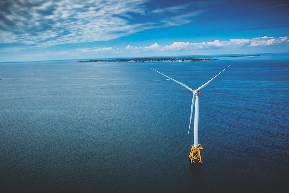 An aerial view of a wind turbine in the ocean with the coast of Block Island and Rhode Island in the background.