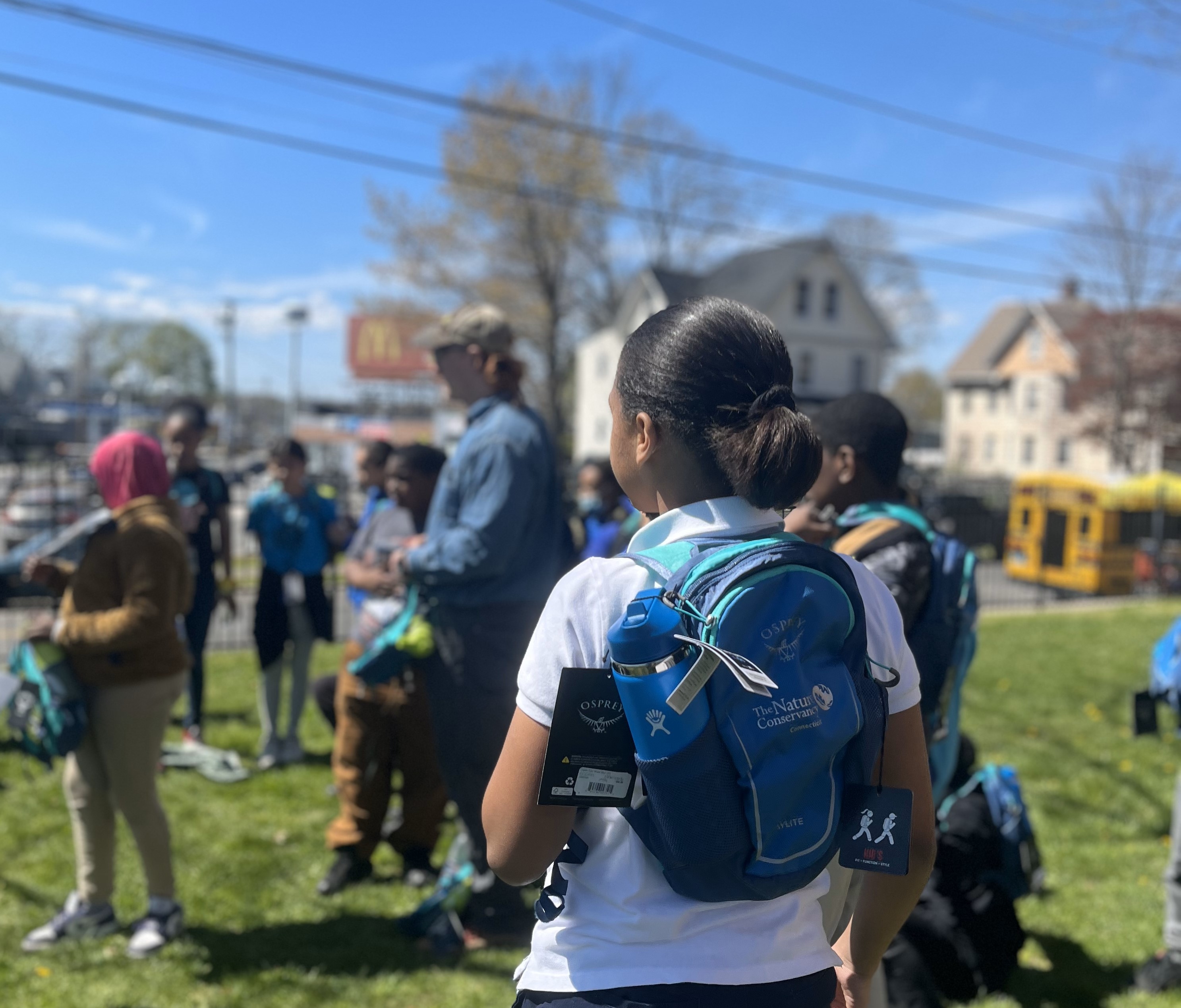 A young girl with a ponytail wears a blue backpack on her back.