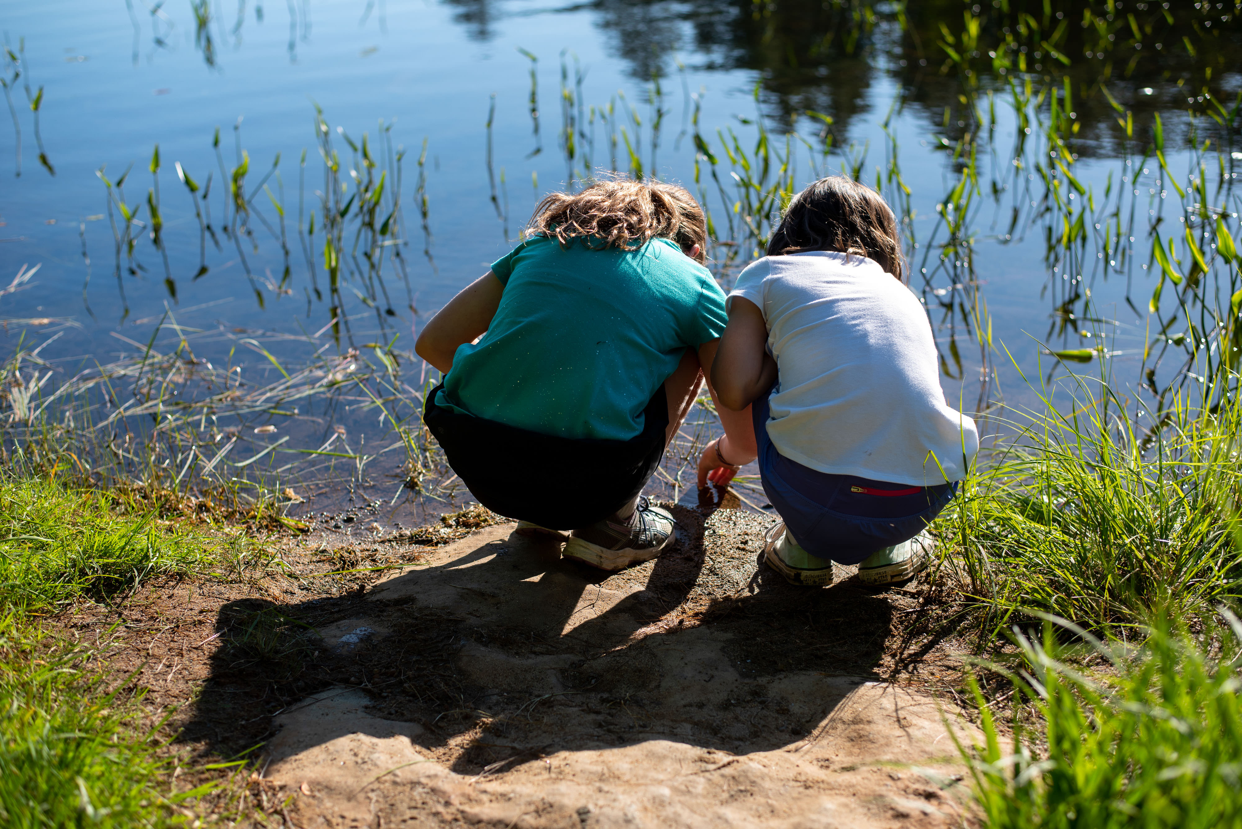 Two children squat down to look into an Adirondacks lake.
