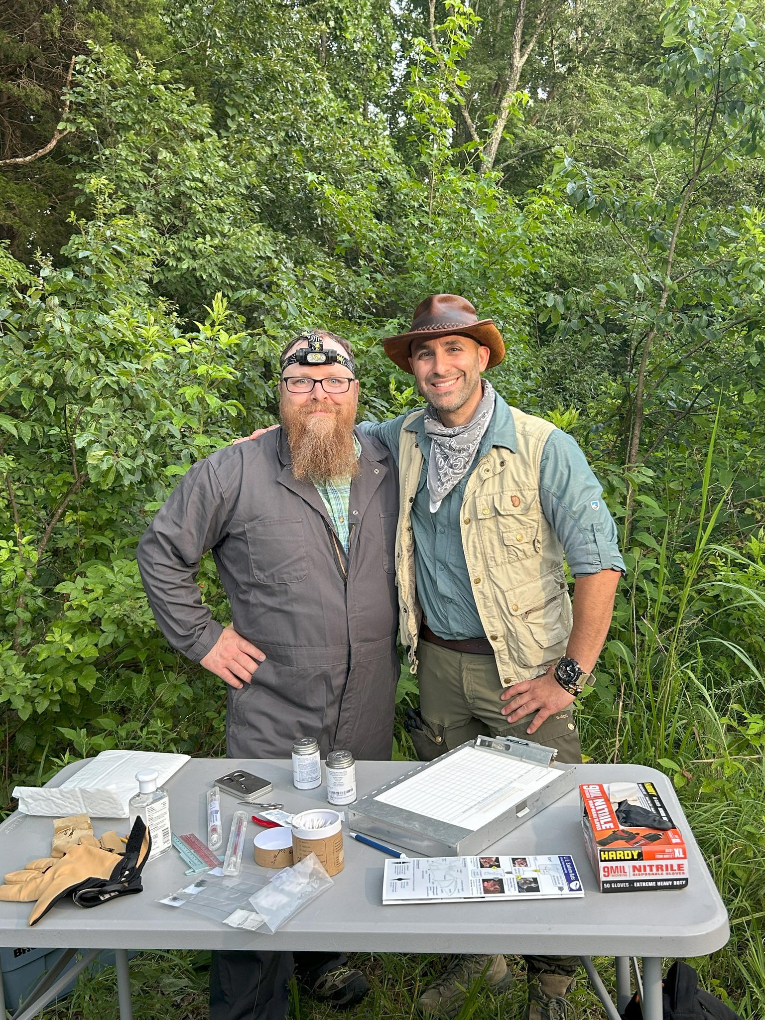 Two men stand behind a table that is set up in a forest.