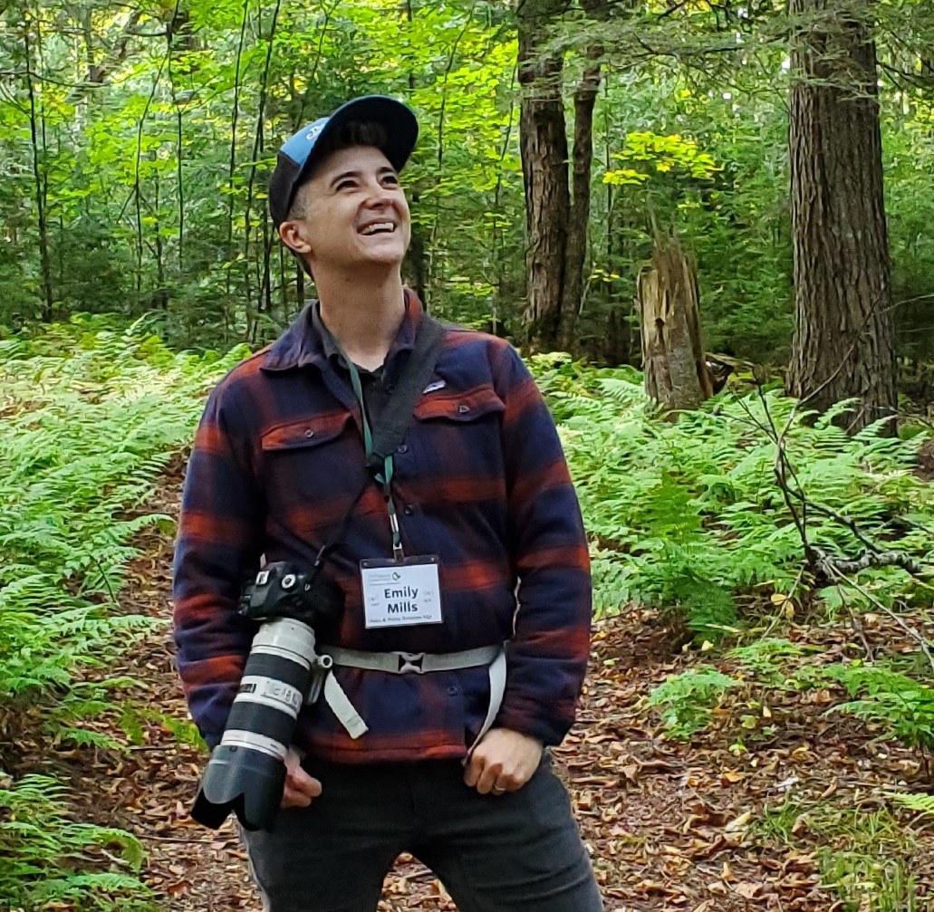 Emily Mills stands in the forest with a camera.