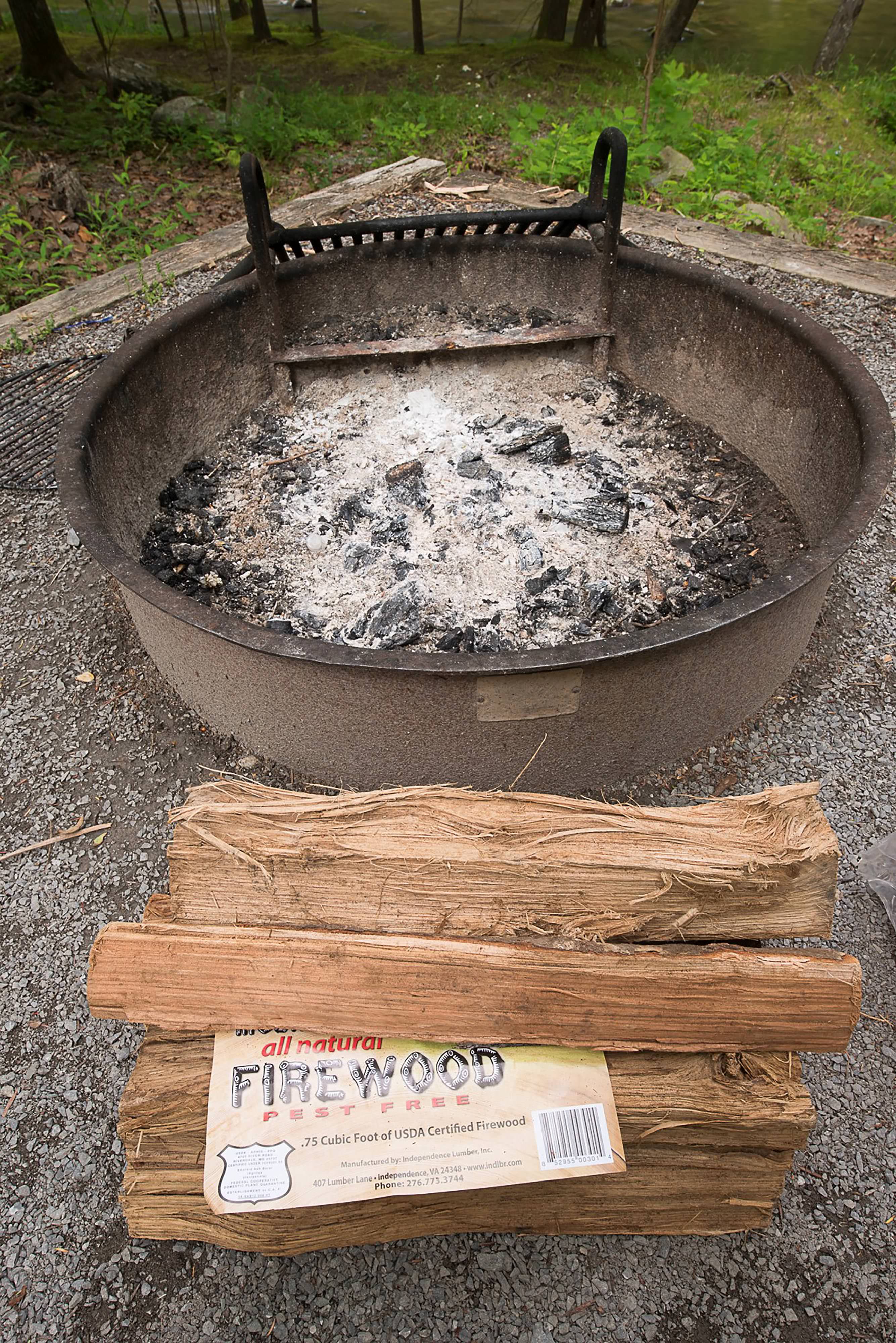 Ash covers the bottom of a large, round metal fire pit. A large piece of firewood lays on the ground in front of the fire pit.
