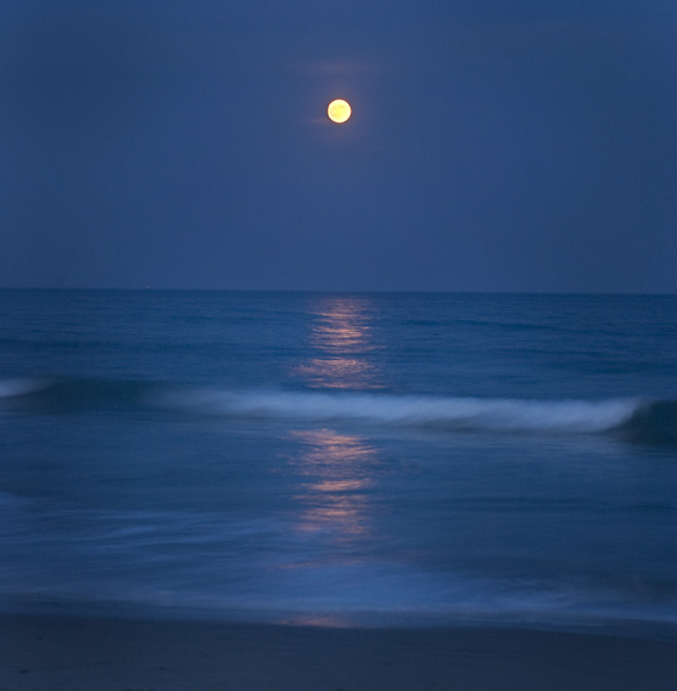 A full moon rises above the ocean, with its light reflected on the waves.