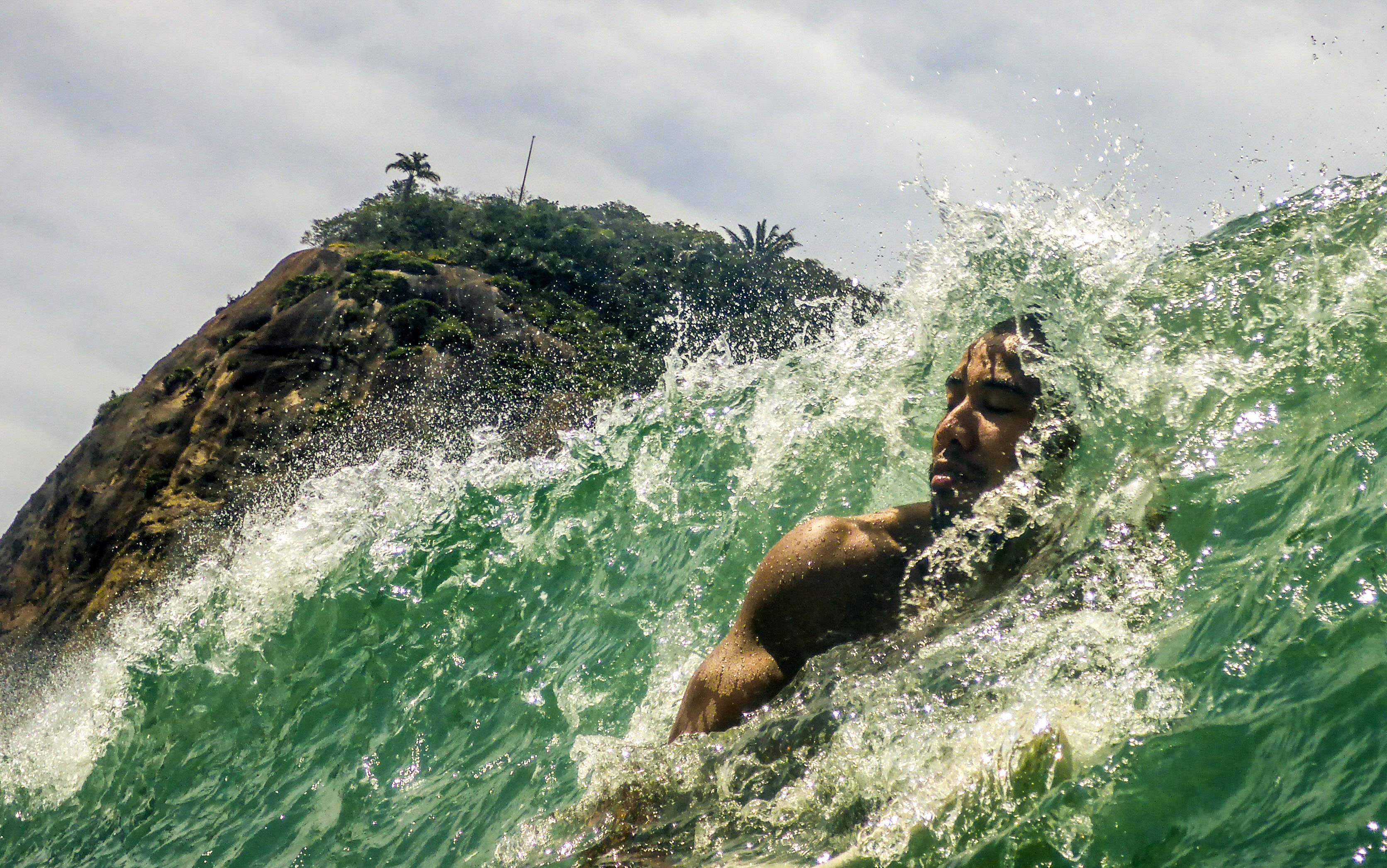 A person swimming through a cresting wave with a green mountain in the background.