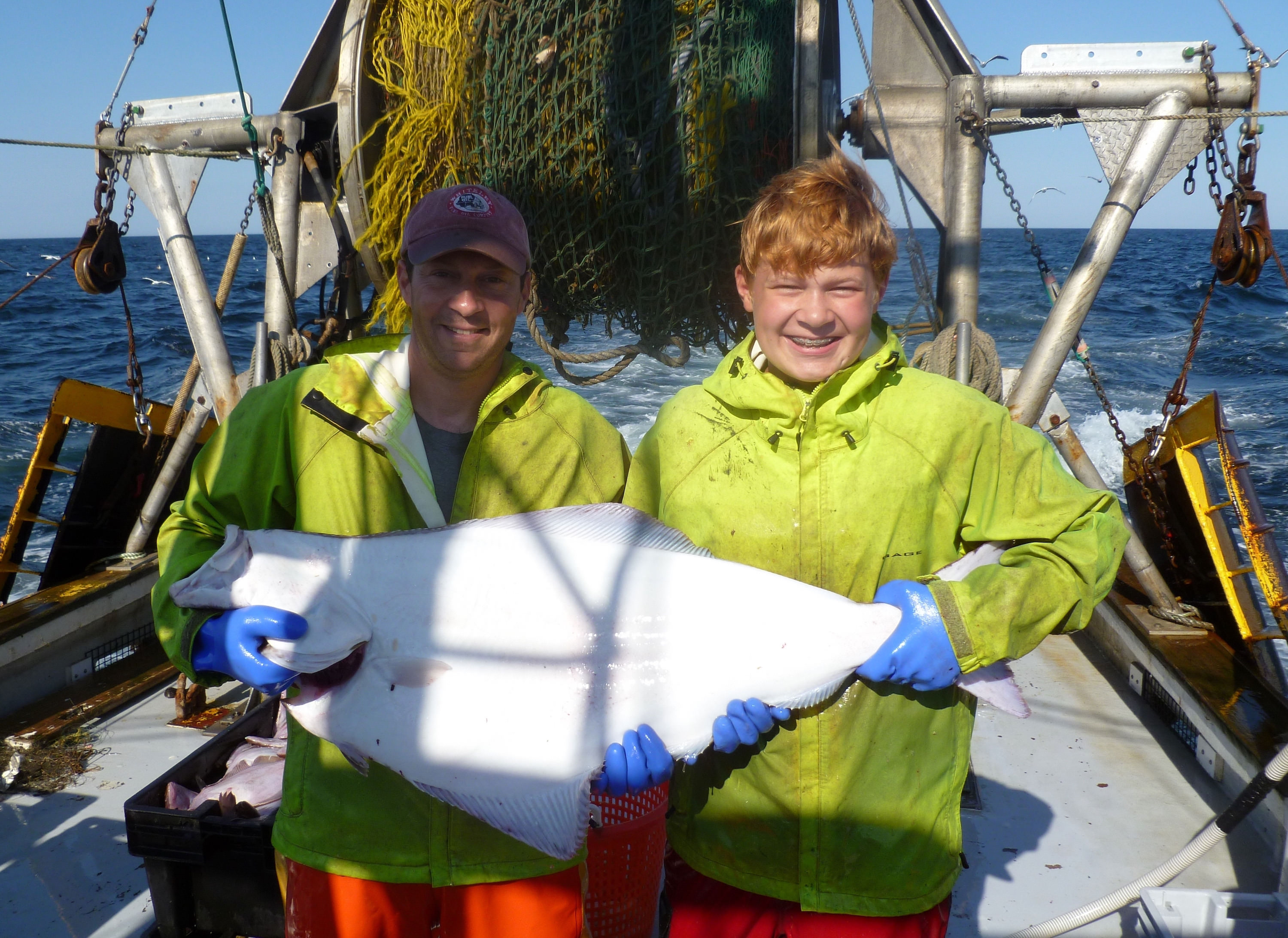 An older man and his son in bright yellow jackets hold a halibut between them, which spans the width of both their chests.