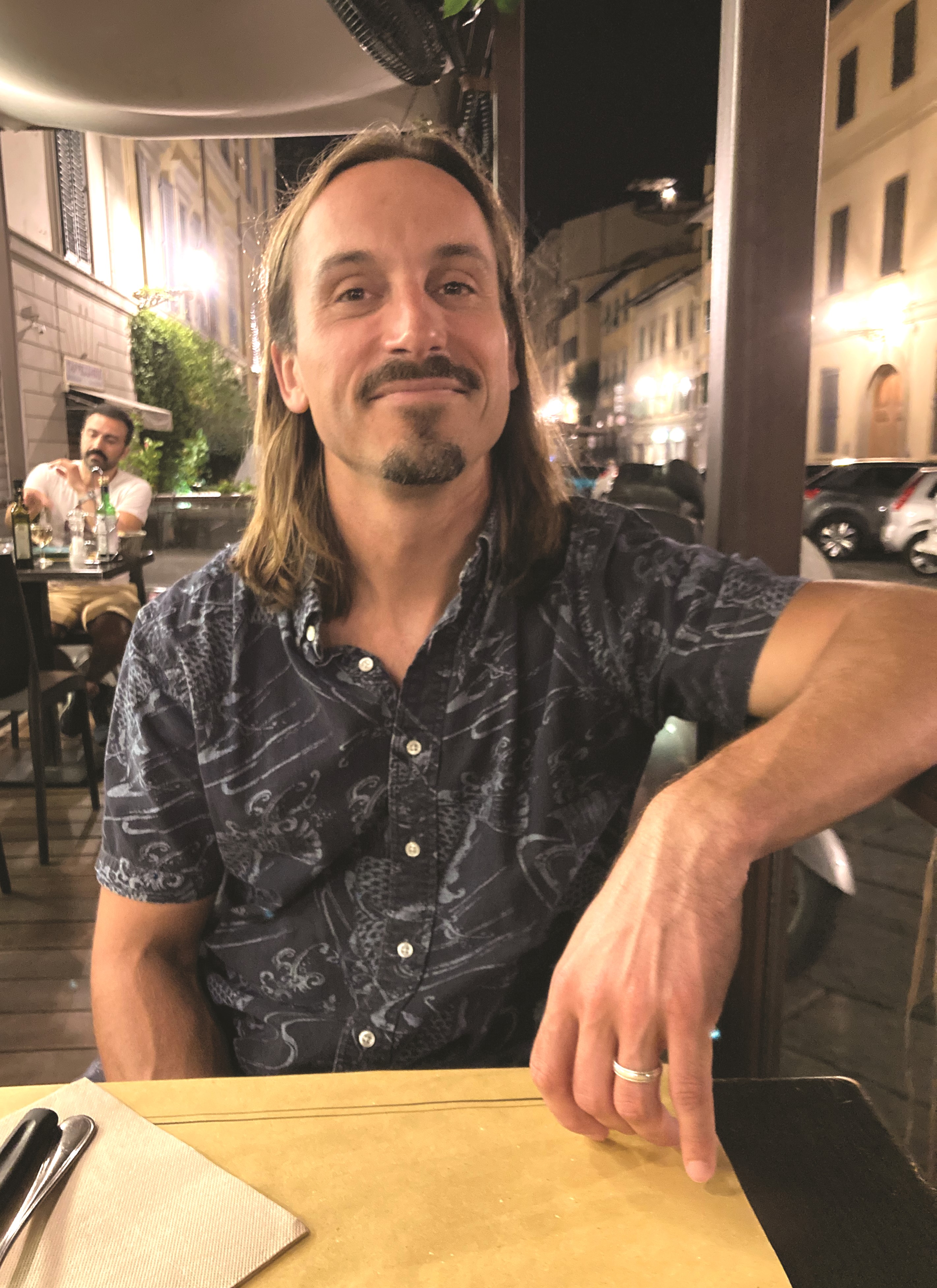 A man in a blue shirt sits at a table outside.