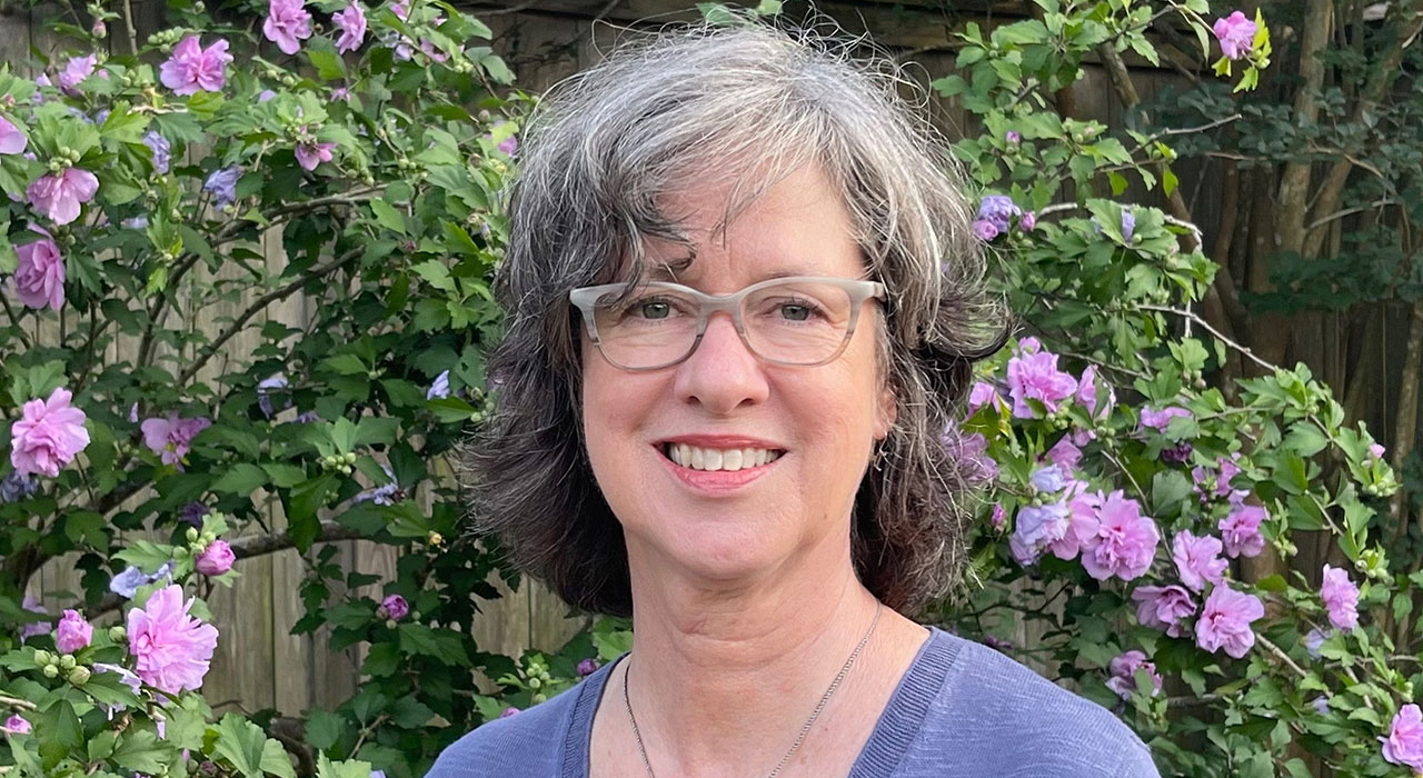 A bush with purple flowers serves as a backdrop for a woman in glasses who is smiling.