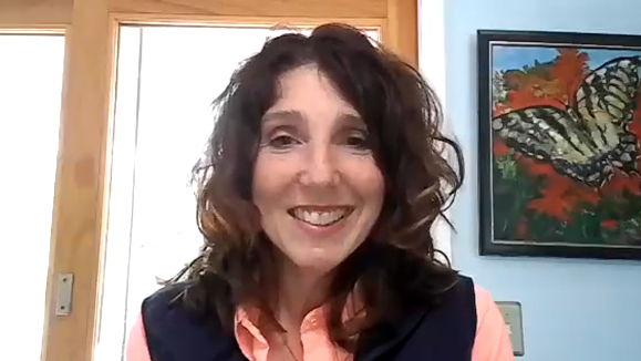 A woman wearing a vest smiles on a computer screen.