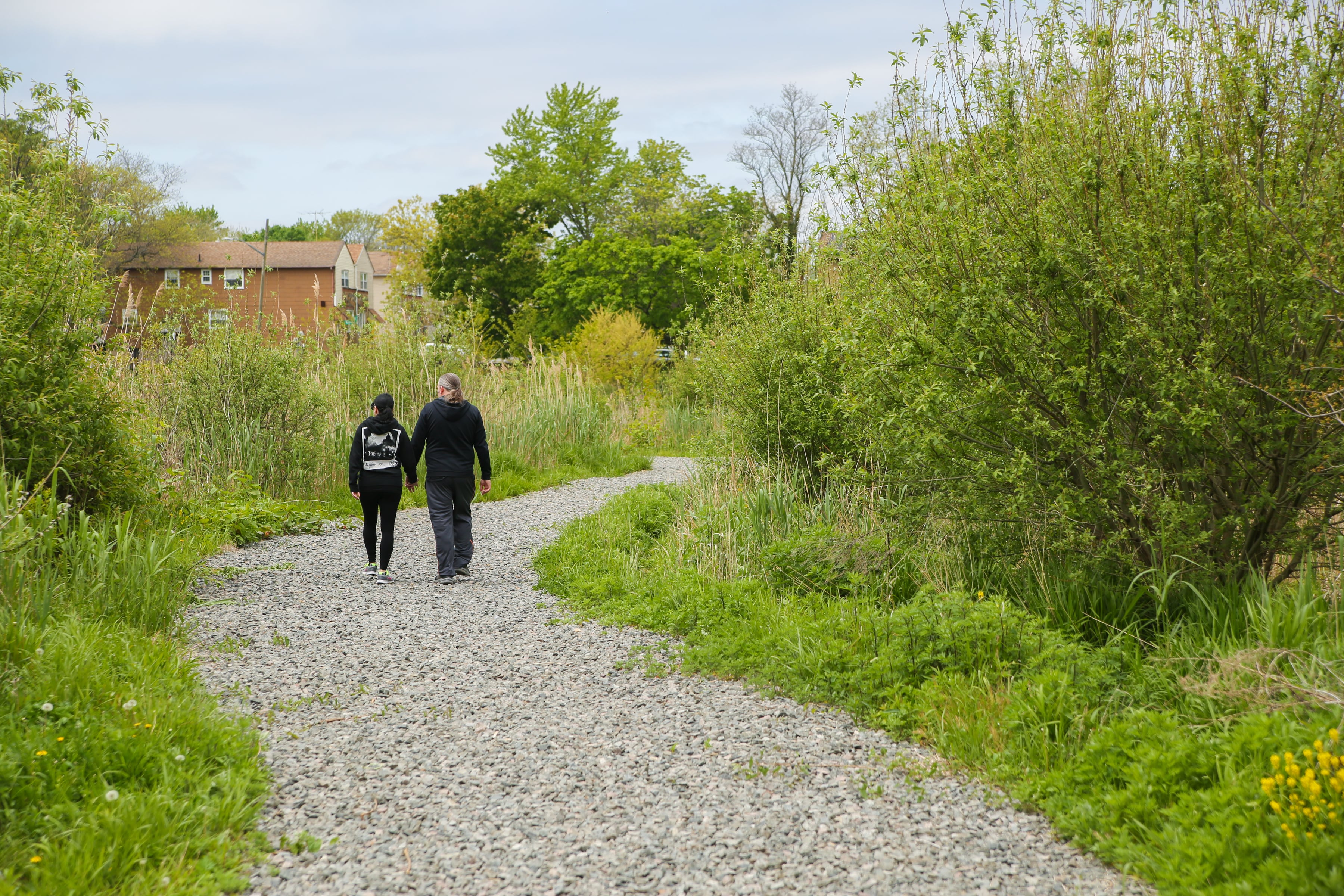 Two people walking away down a graveled path in a wooded section of a neighborhood with the roof of a house emerging in the distance.