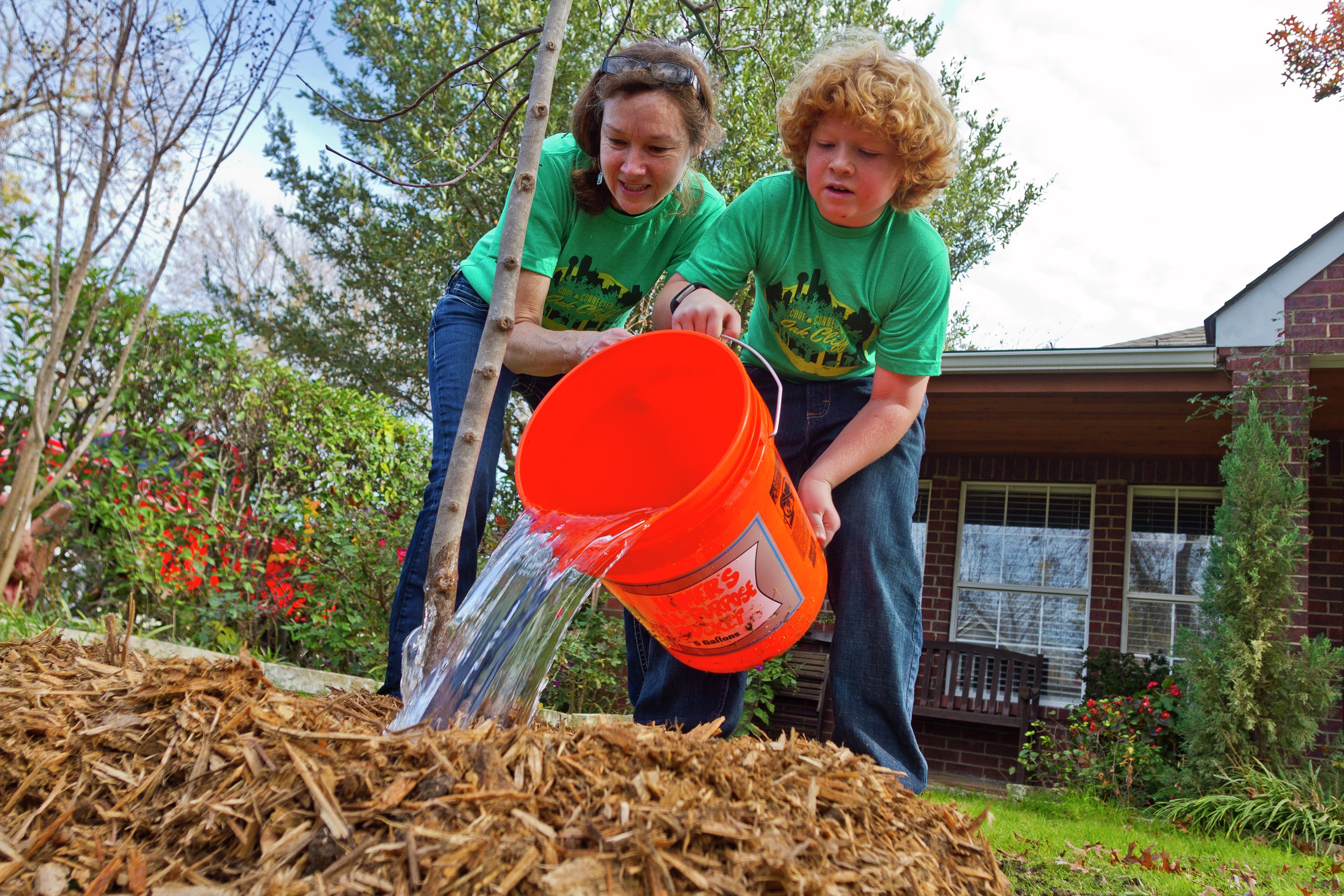 Two people use a large orange bucket to water a tree.