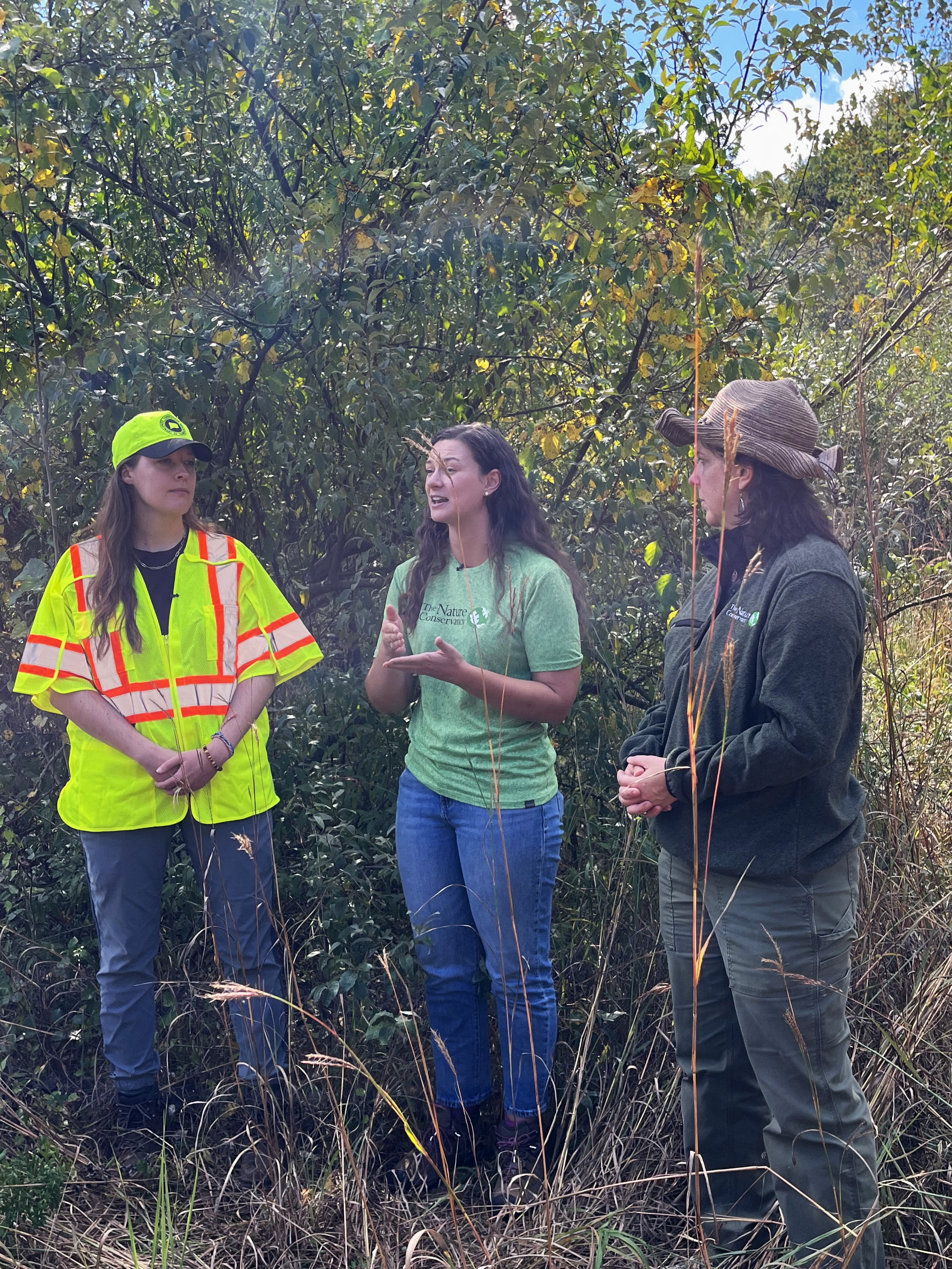 Three women have a conversation against a forest backdrop.